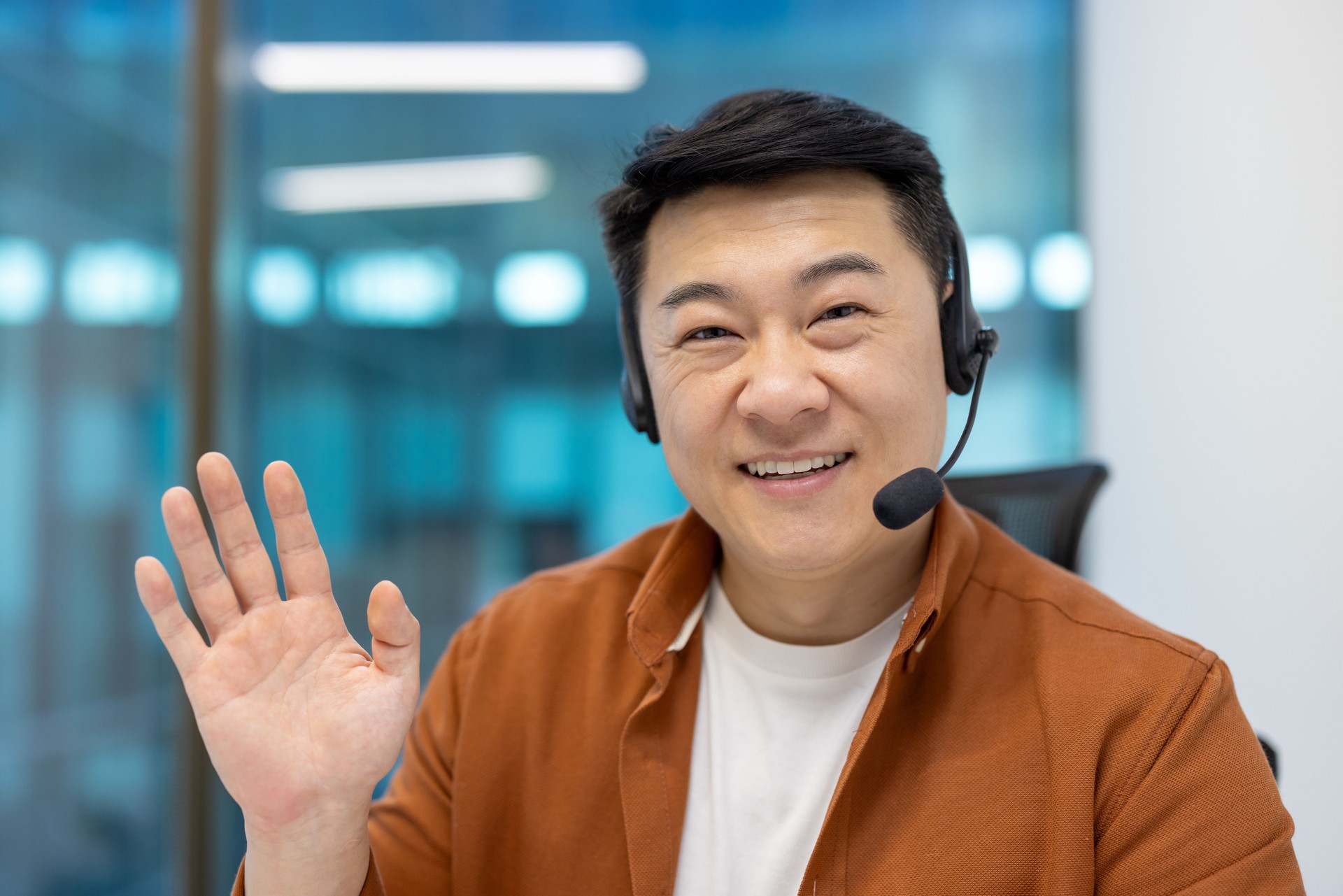 Smiling asian businessman wearing headset waving hand during video conference in modern office setting
